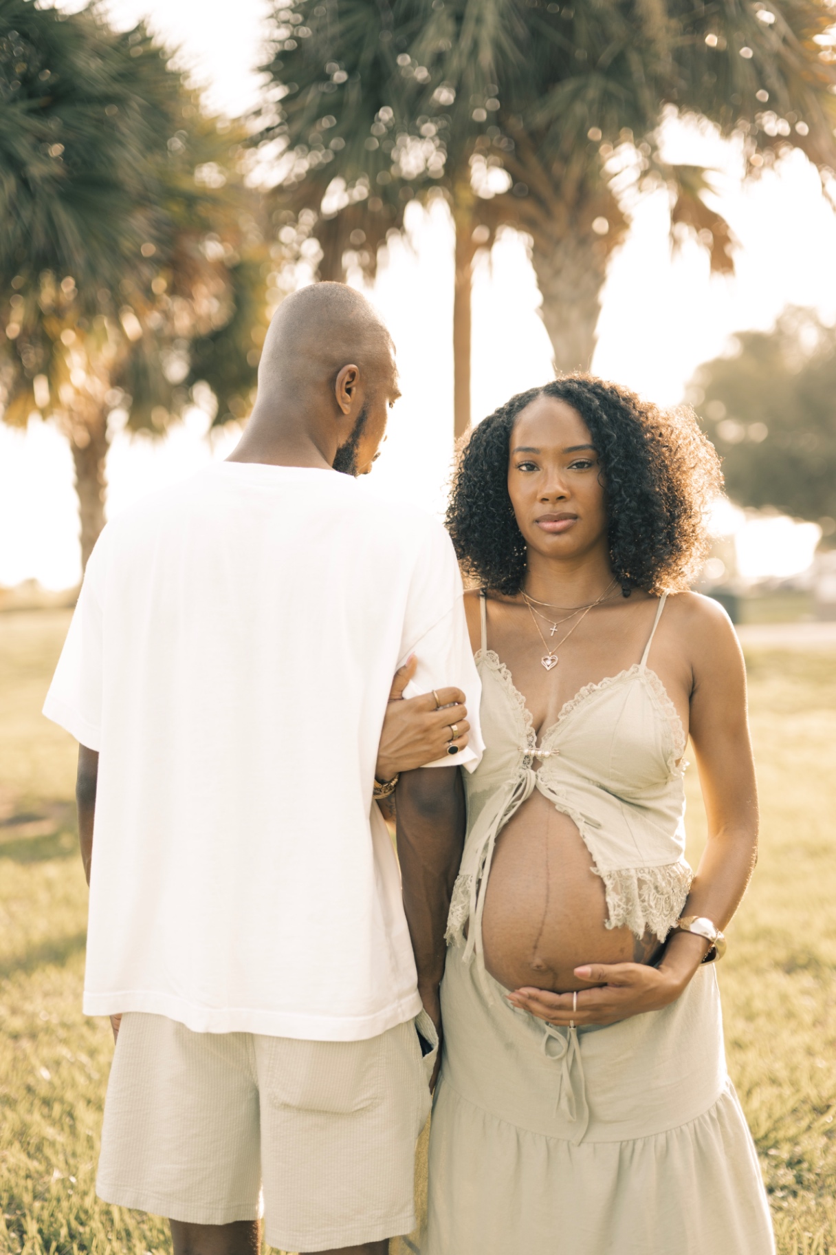 Expecting mother and partner standing beneath palm trees at golden hour